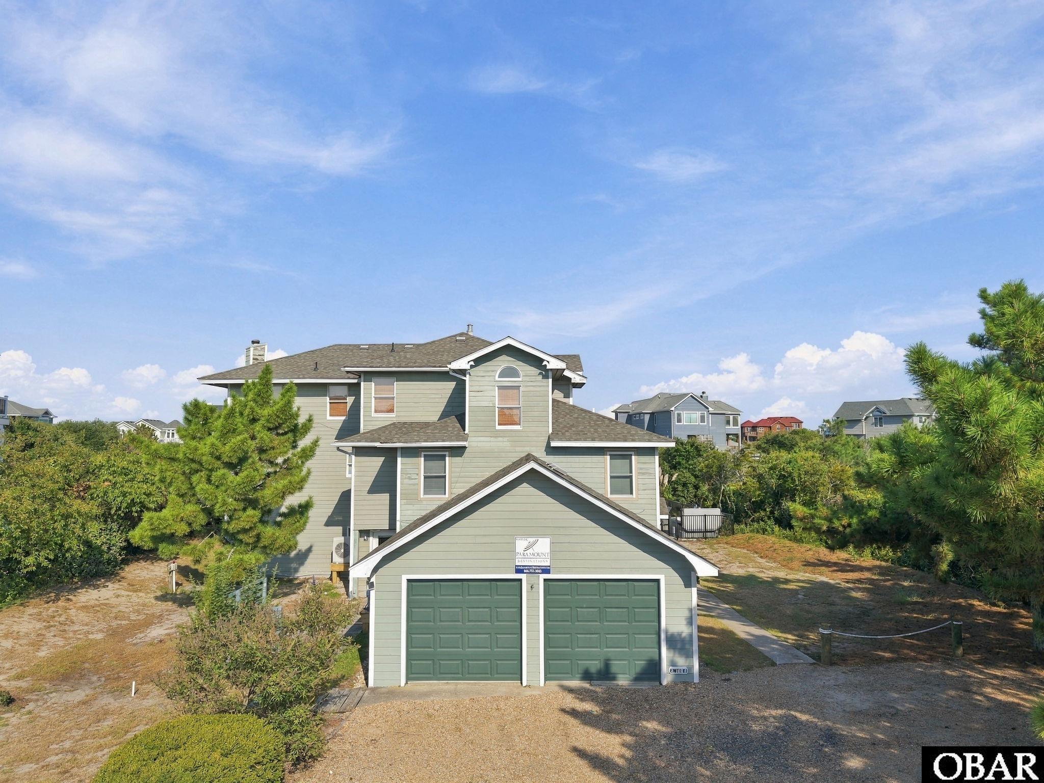 Sage-green coastal house with two green garage doors, framed by pine trees and shrubs.
