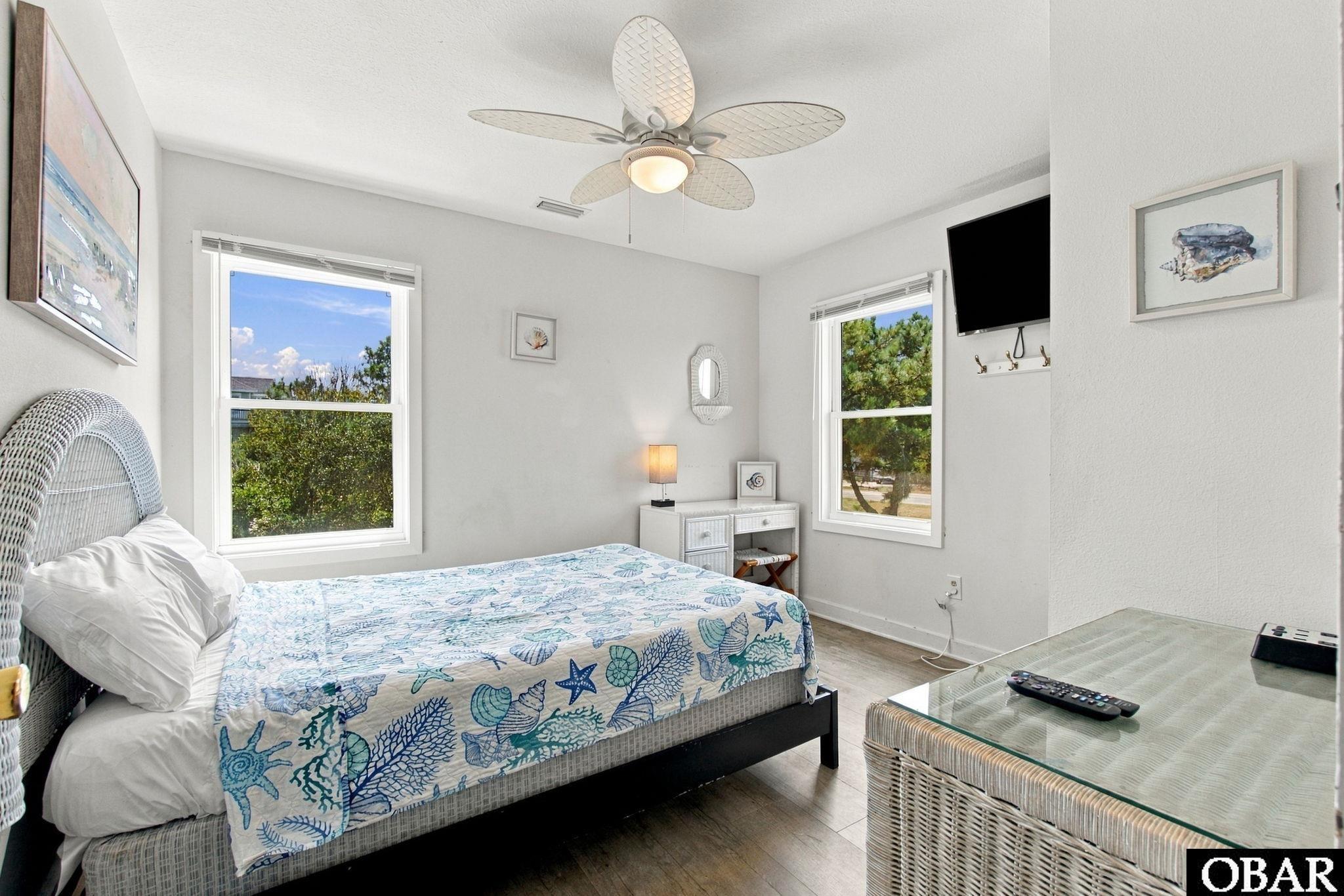 Furnished bedroom with seashell-patterned bedding, wicker headboard, two windows, and a ceiling fan.