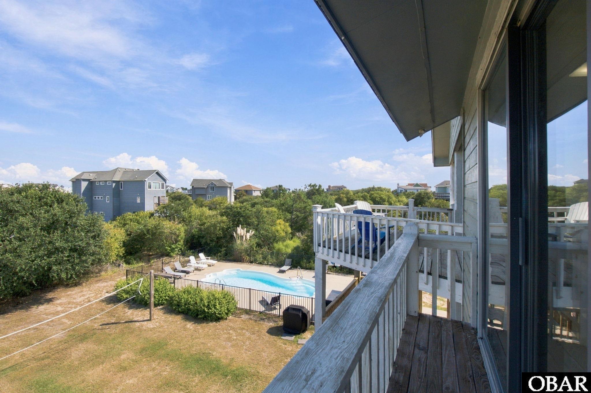 Raised deck overlooks a fenced swimming pool and neighboring homes beyond dense greenery.