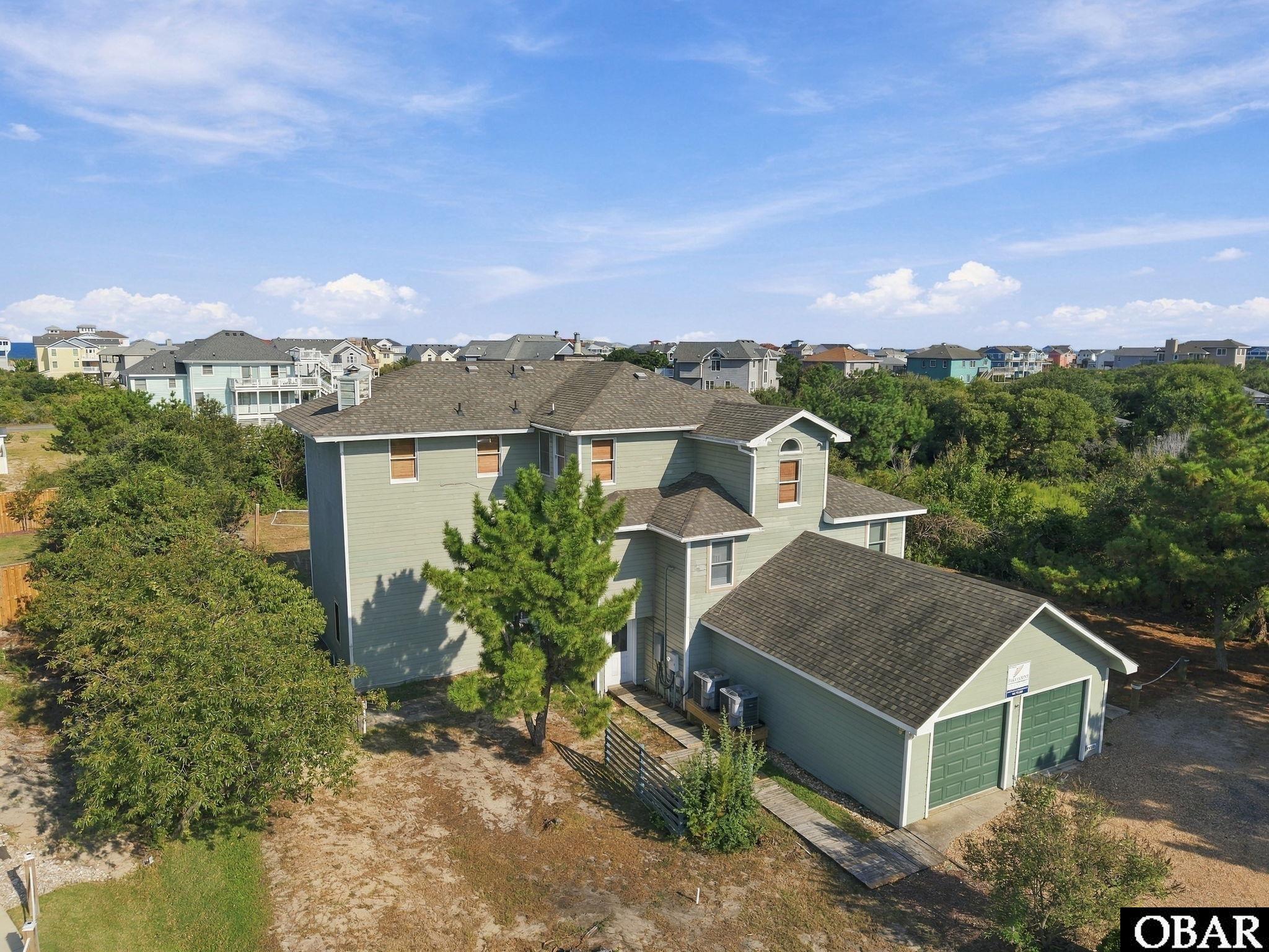 Aerial view of a sage-green home with a detached garage, surrounded by trees and neighboring houses.