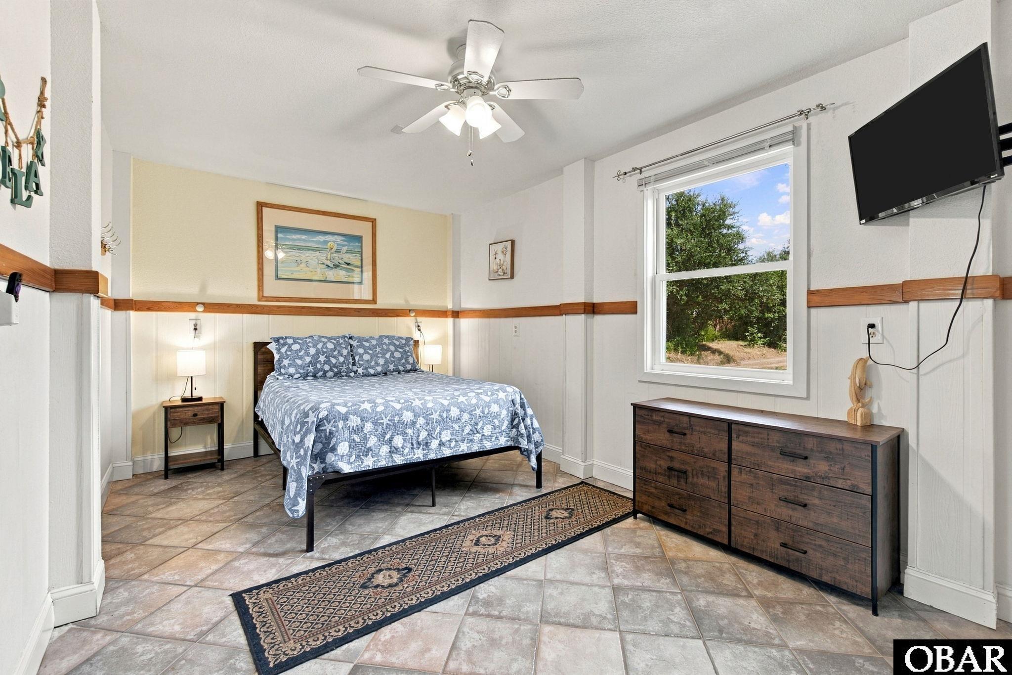 Bedroom with a blue patterned bed, tiled floor, dark wood dresser, and window overlooking trees.