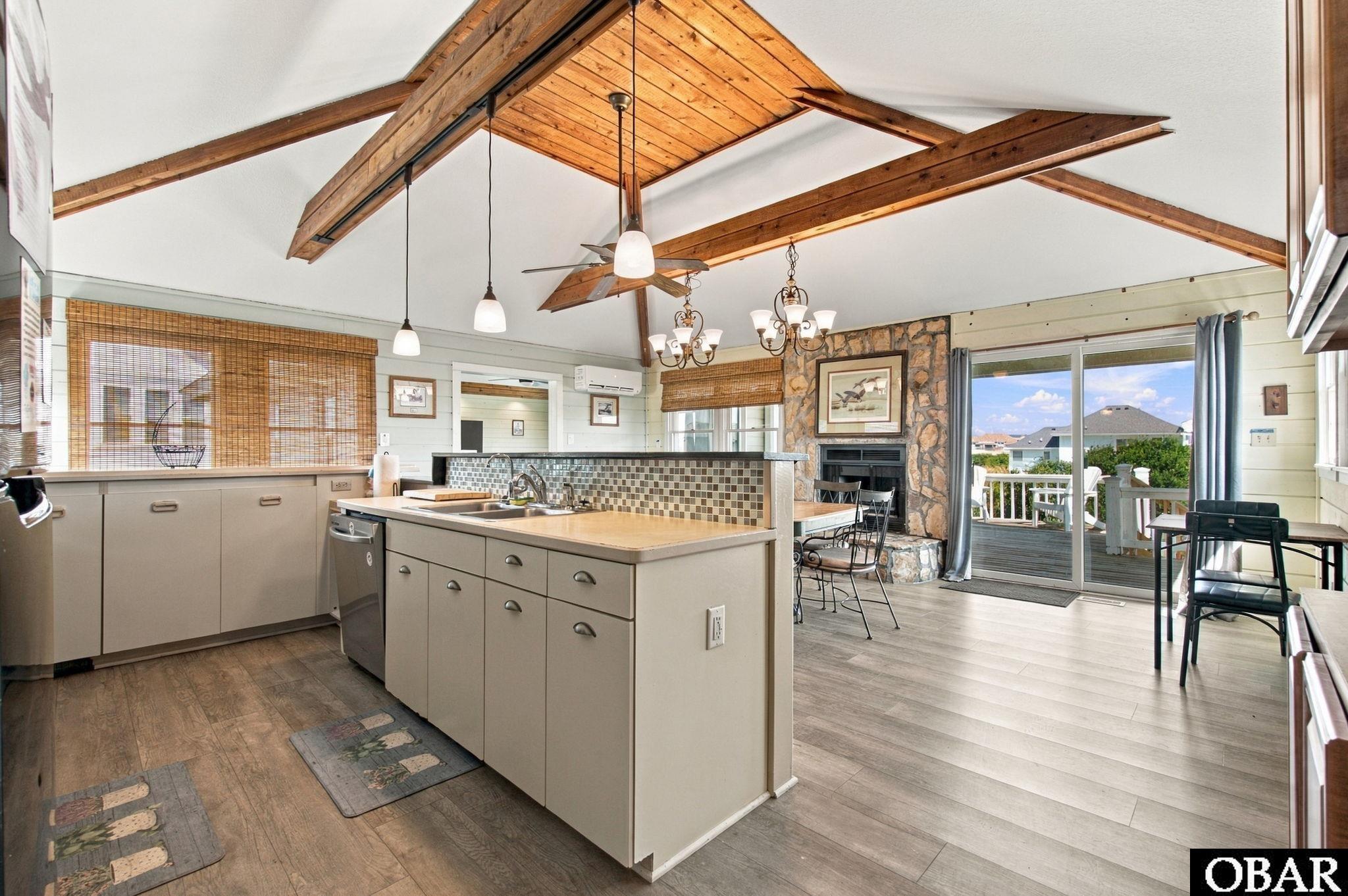 Open-plan kitchen with central island, vaulted timber-beam ceiling, stone fireplace, and sliding doors to a deck.