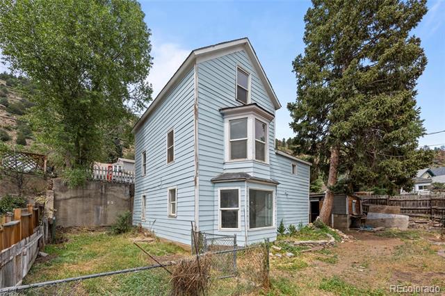 Two-story light-blue house with bay windows, chain-link fence and mature trees.
