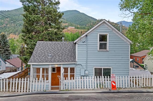 Light-blue house with a steep gabled upper story and white picket fence, backed by forested mountains.