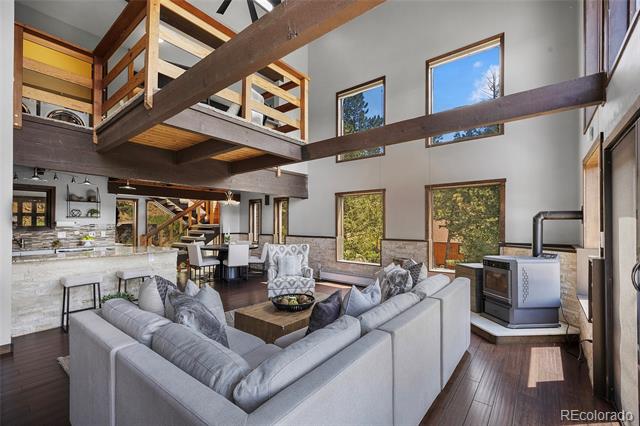 Living room with a gray sectional, exposed beams, an open loft, and large windows overlooking trees.