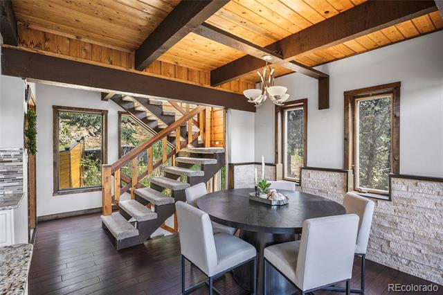 Dining area with a round table, exposed wood beams, stone accents, and a staircase beside tall windows.