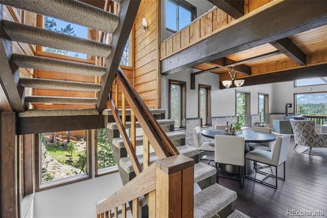 Open living and dining area with a carpeted staircase, exposed beams, and large windows.