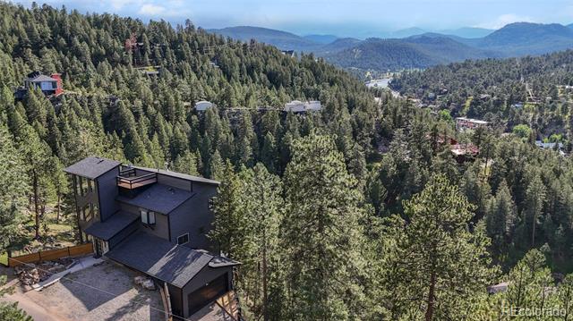 Aerial view of a dark modern home among evergreen trees, with a mountain valley beyond.