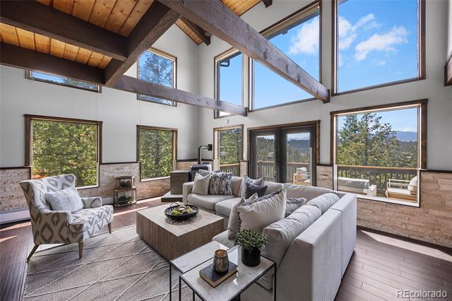 Vaulted living room with exposed timber beams, large windows, and a gray sectional facing mountain views.