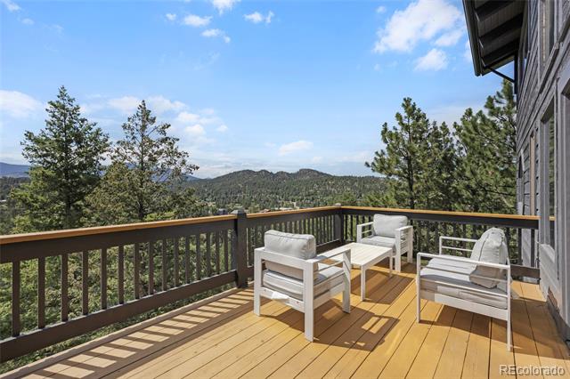 Furnished wooden deck overlooking pine-covered mountain ridges, with white outdoor seating.