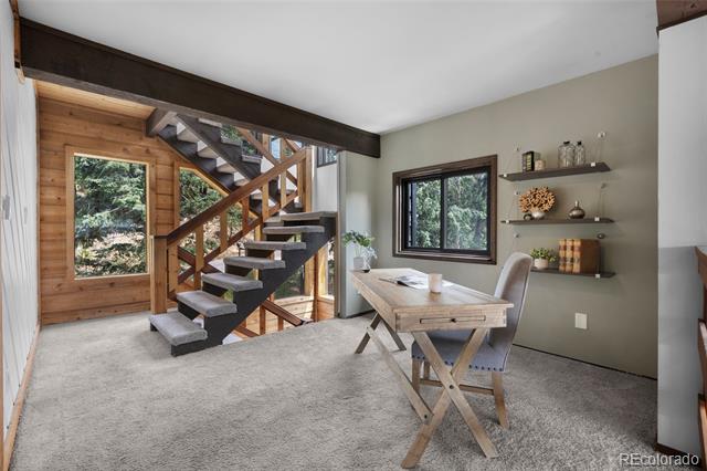 Workspace beside an open staircase, with a wood table, floating shelves, and windows overlooking trees.