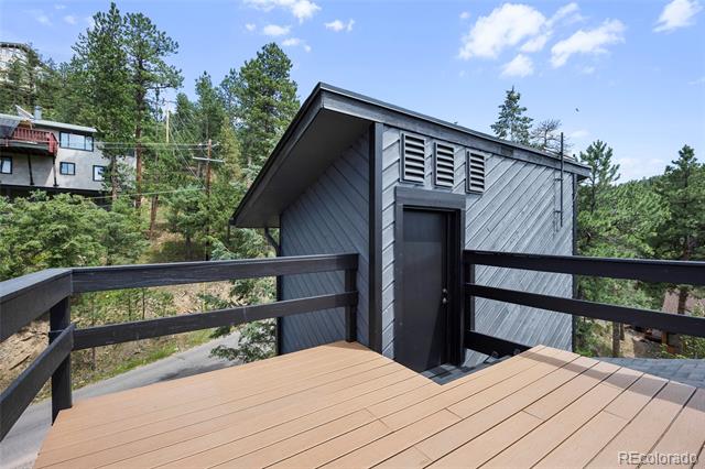 Elevated wood deck beside a gray-sided structure, surrounded by evergreen trees.
