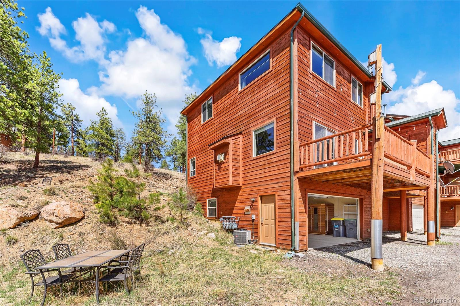 Wood-sided home with a raised deck and open garage beside a rocky, pine-covered hillside.