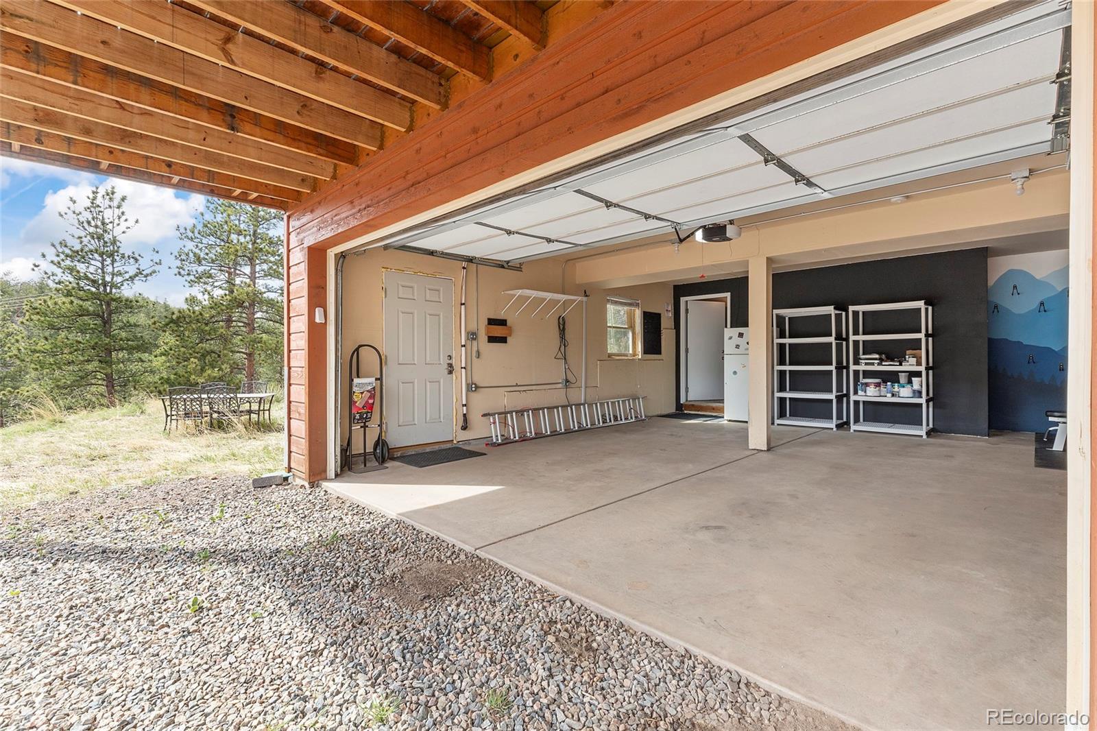 Open garage with storage shelves and exposed wood ceiling, beside a gravel area and pine trees.