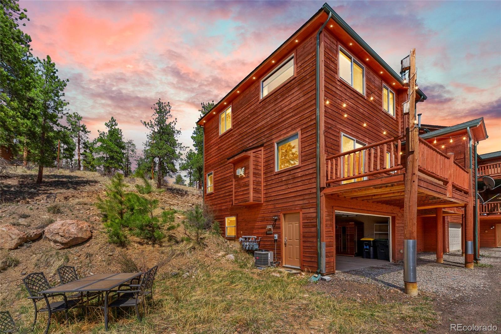 Red wood-sided multi-story home with a raised deck beside a pine-covered hillside at dusk.