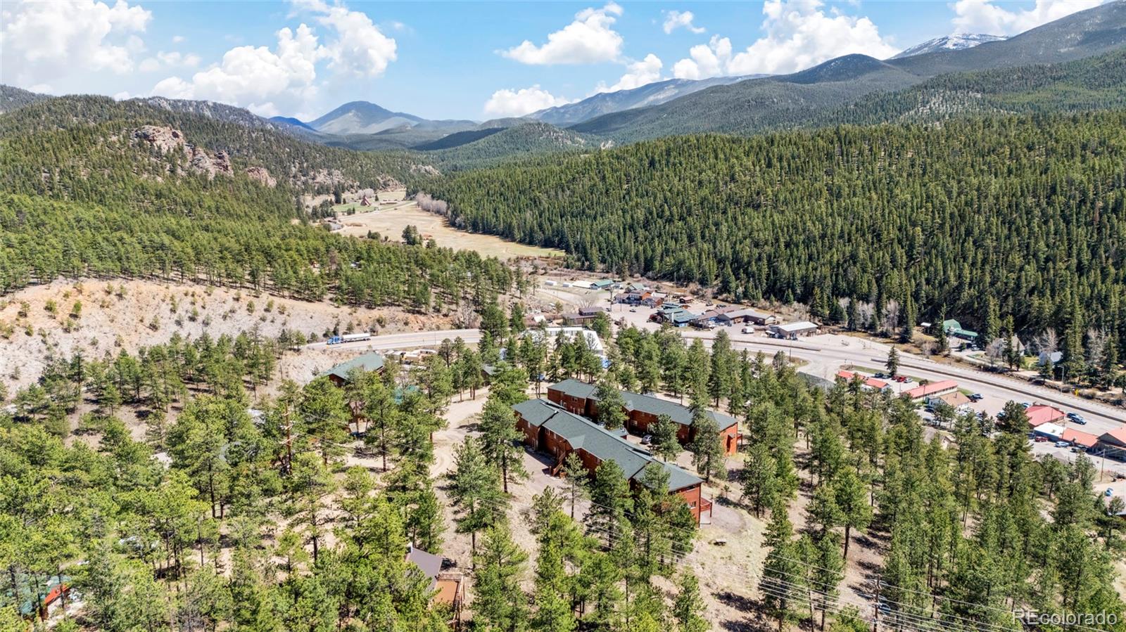 Aerial view of mountain buildings among pine trees, with a highway and forested peaks beyond.