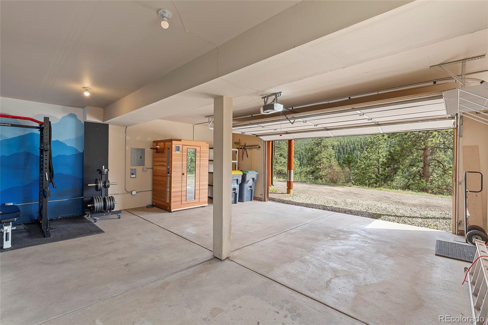 Garage with a home gym and wooden sauna beside an open door overlooking pine trees.