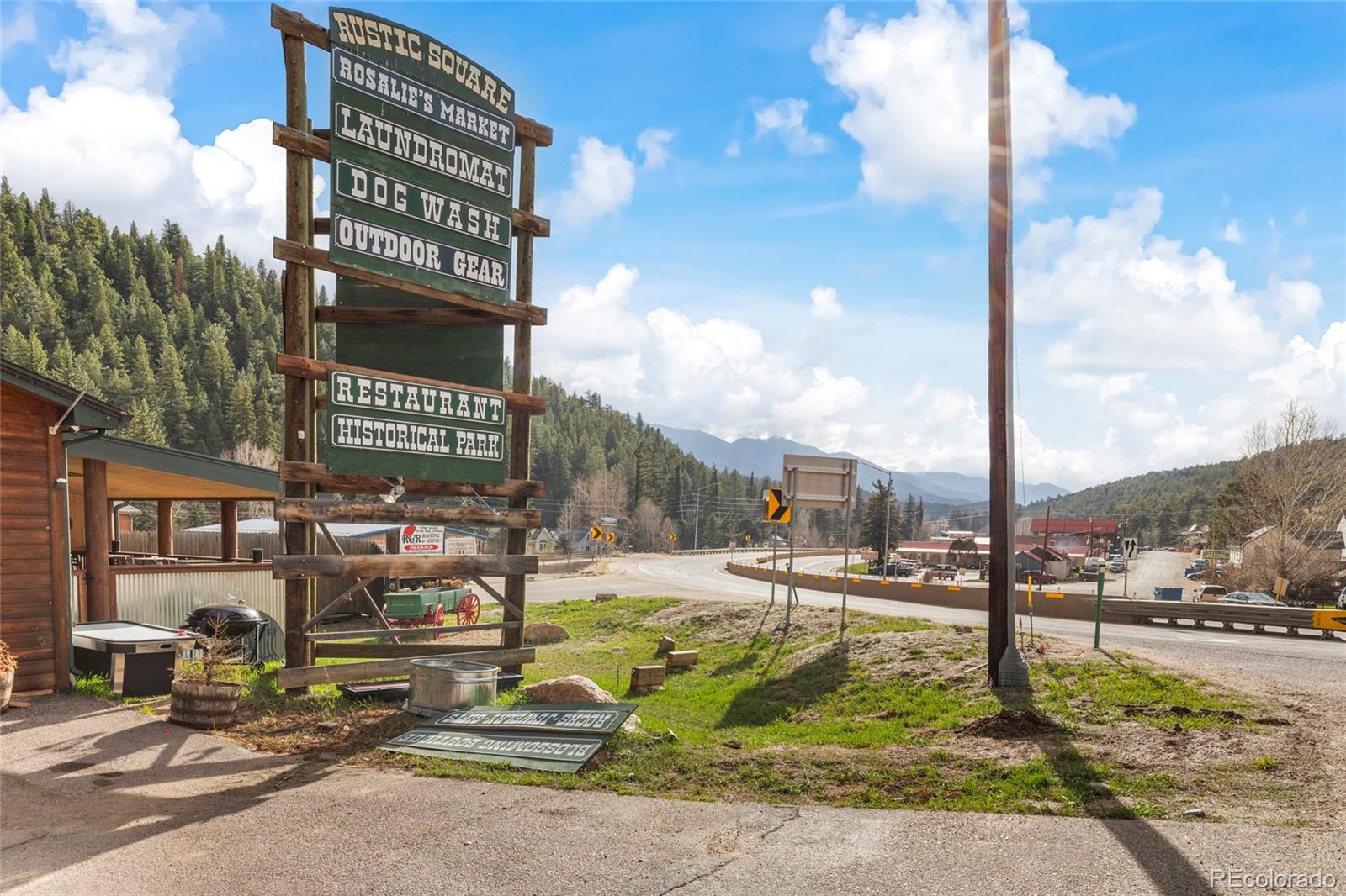 Roadside Rustic Square sign lists shops and services beside a highway in a forested mountain community.