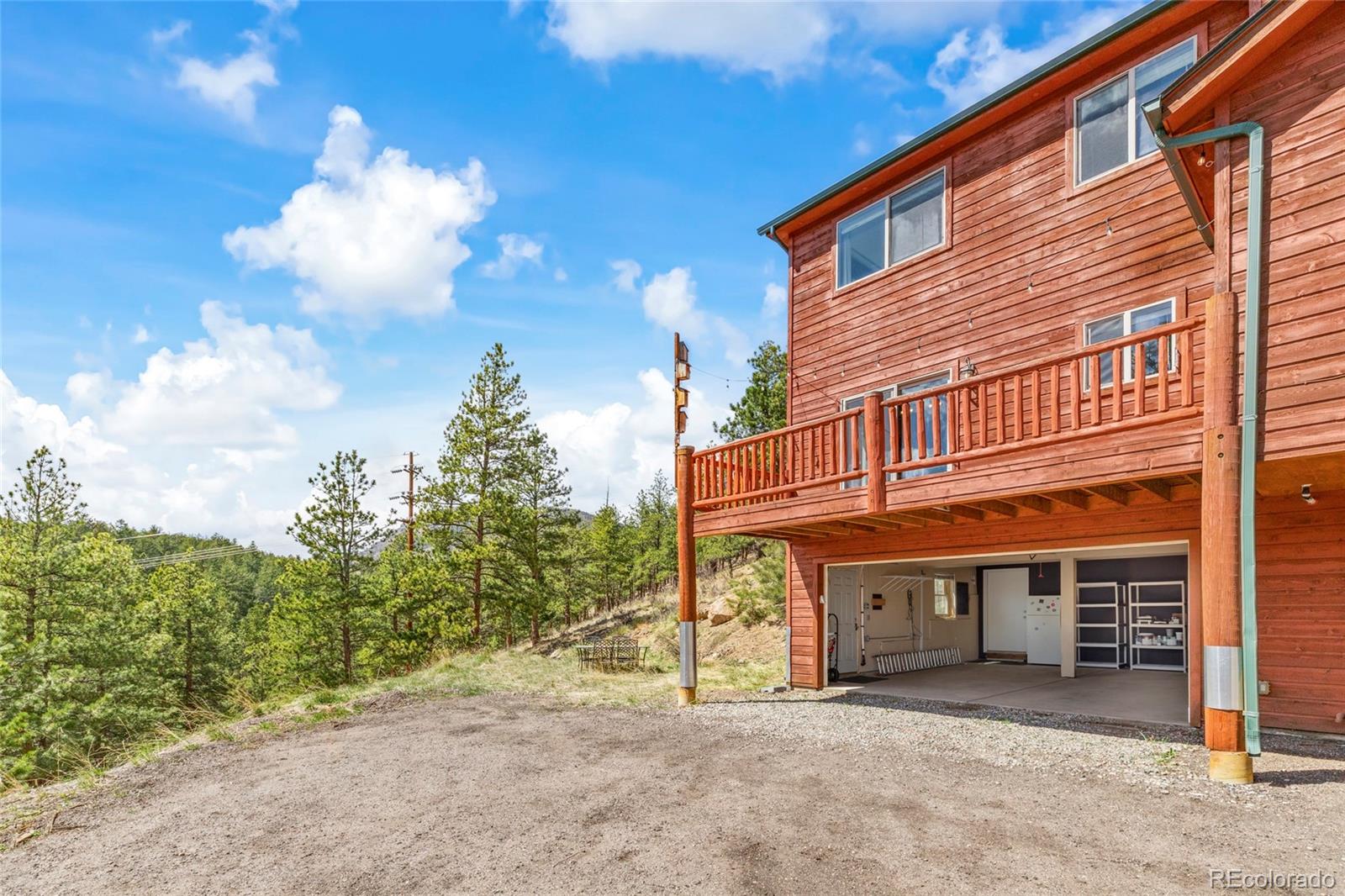 Wood-sided home with a raised deck and open garage beside a wooded hillside.