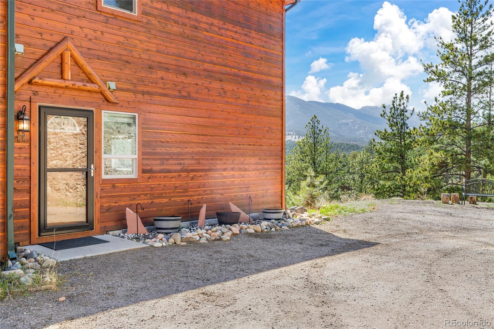 Wood-sided mountain home with a glass entry door, gravel drive, pine trees, and mountain views.