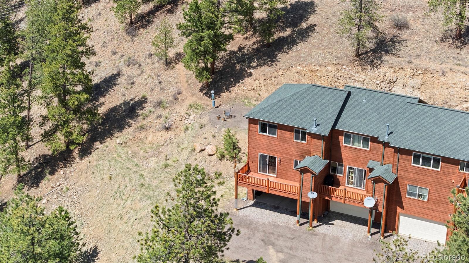 Aerial view of a wood-sided residence with decks and covered parking on a pine-covered hillside.