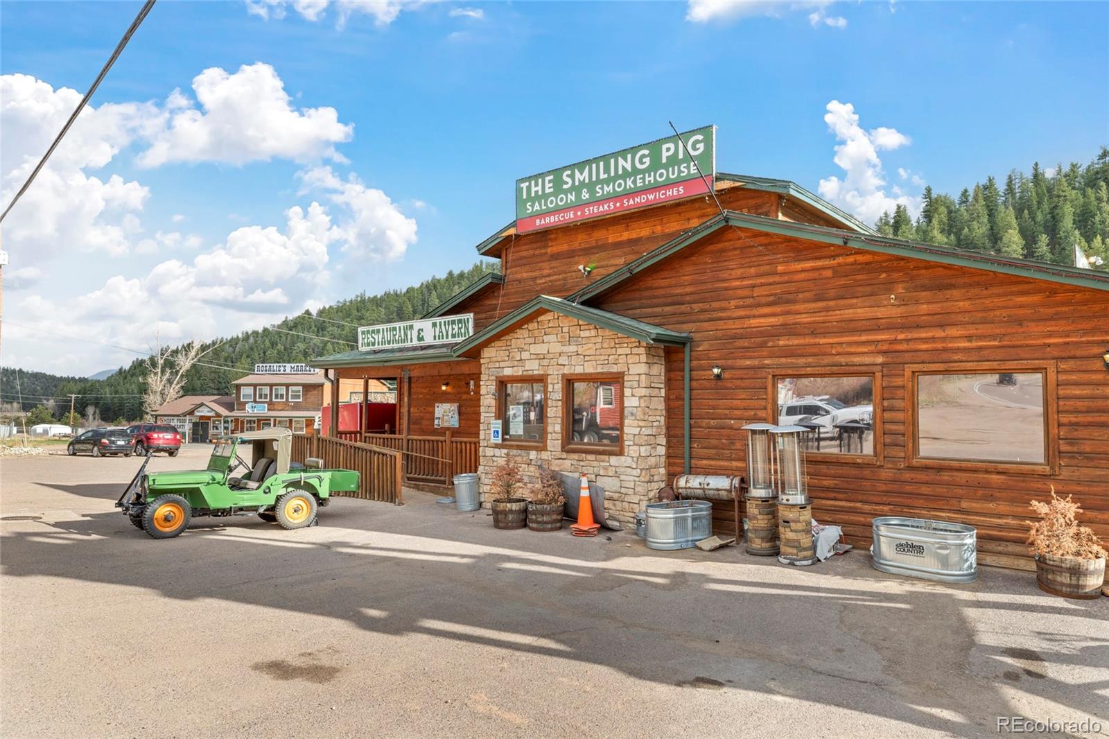 Rustic wood-and-stone restaurant with The Smiling Pig Saloon & Smokehouse sign and a green jeep outside.