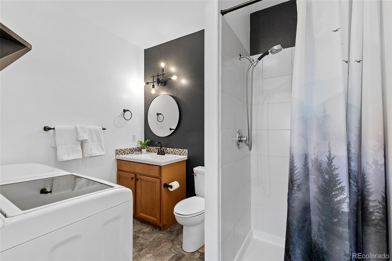 Bathroom with a tiled shower, forest-print curtain, wooden vanity, and washer and dryer.