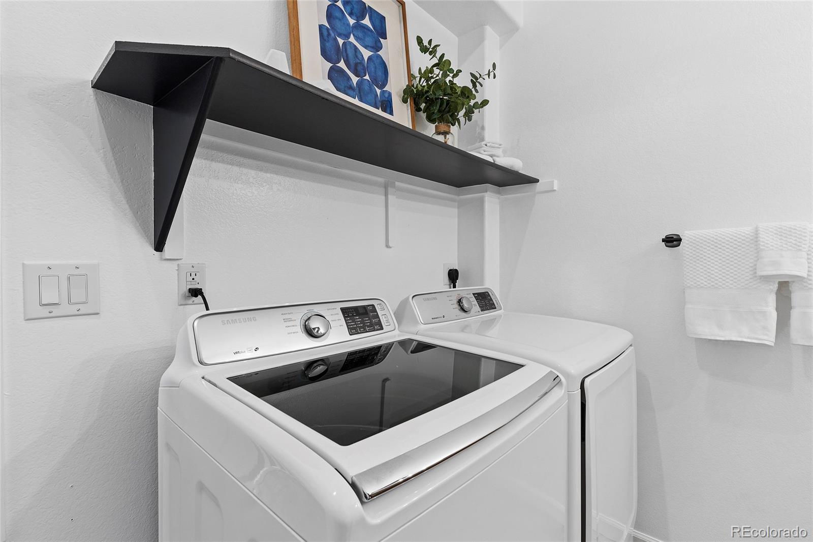 Laundry room with a white top-loading washer and dryer beneath a black shelf.