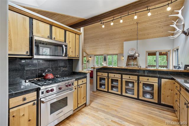 Rustic kitchen with light wood cabinetry, black counters, stainless steel range, and glass-front island.