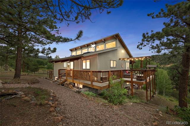 Two-story gray home with a wraparound wooden deck among pine trees at dusk.
