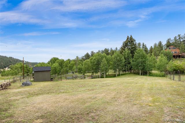 Grassy yard with a small shed, fencing, and trees; a house sits on the hillside in the distance.