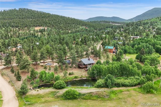 Aerial view of a mountain home beside a pond, surrounded by pine forest and distant hills.