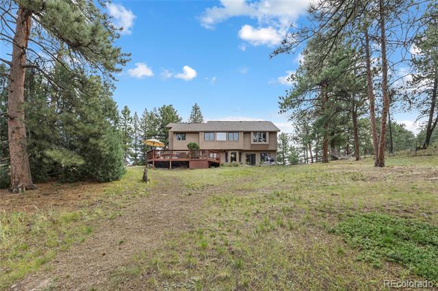 Two-story brown house with a deck on a sloping, pine-dotted lot.