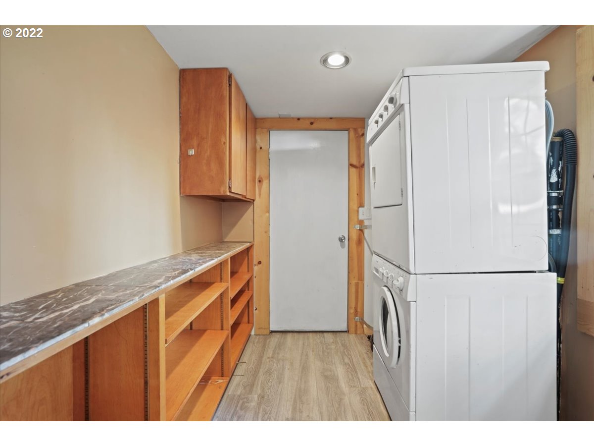 Laundry room with stacked washer and dryer beside a long counter and open wooden shelving.