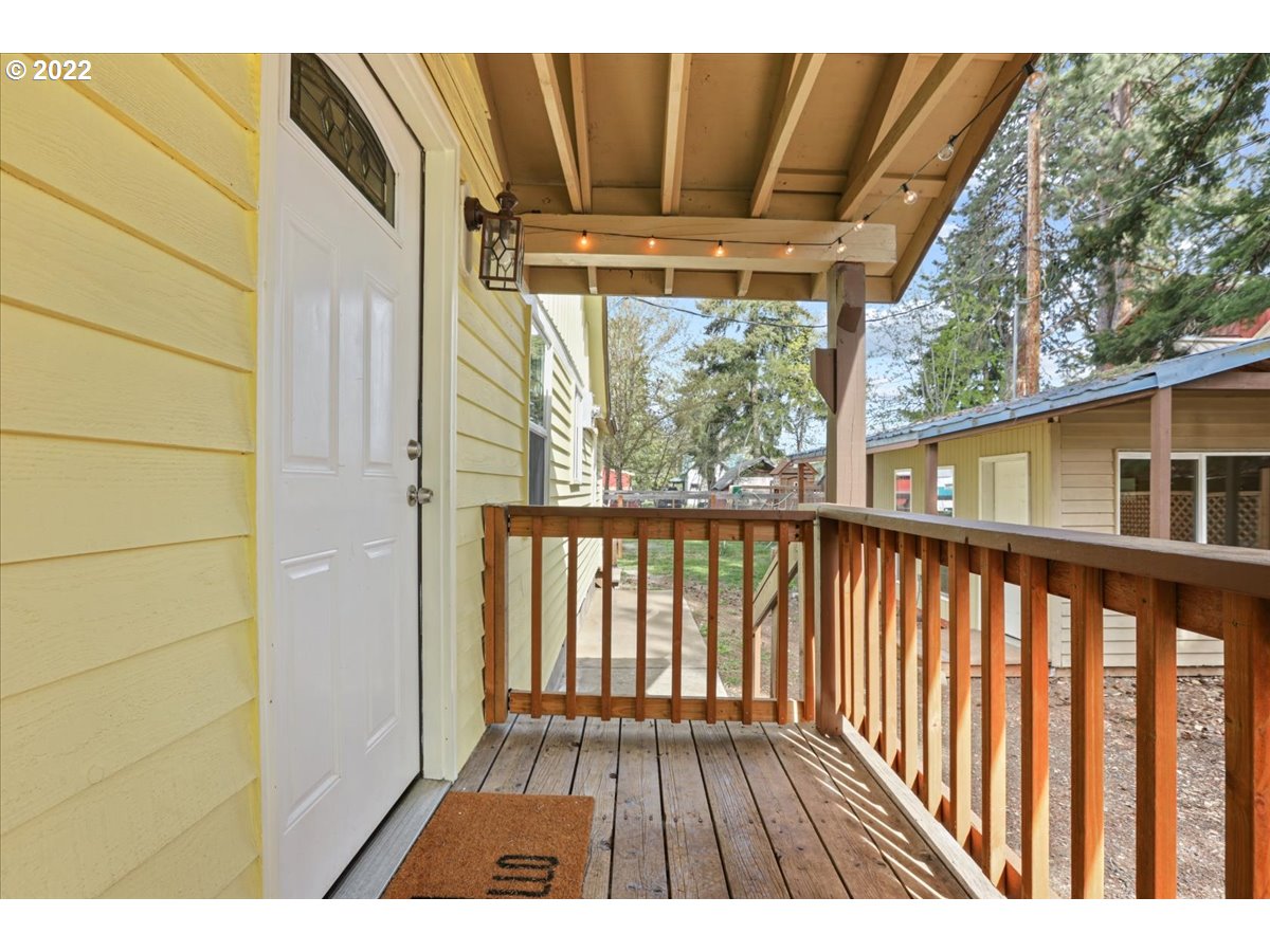 Covered wooden porch with brown railings, exposed beams and a white entry door.