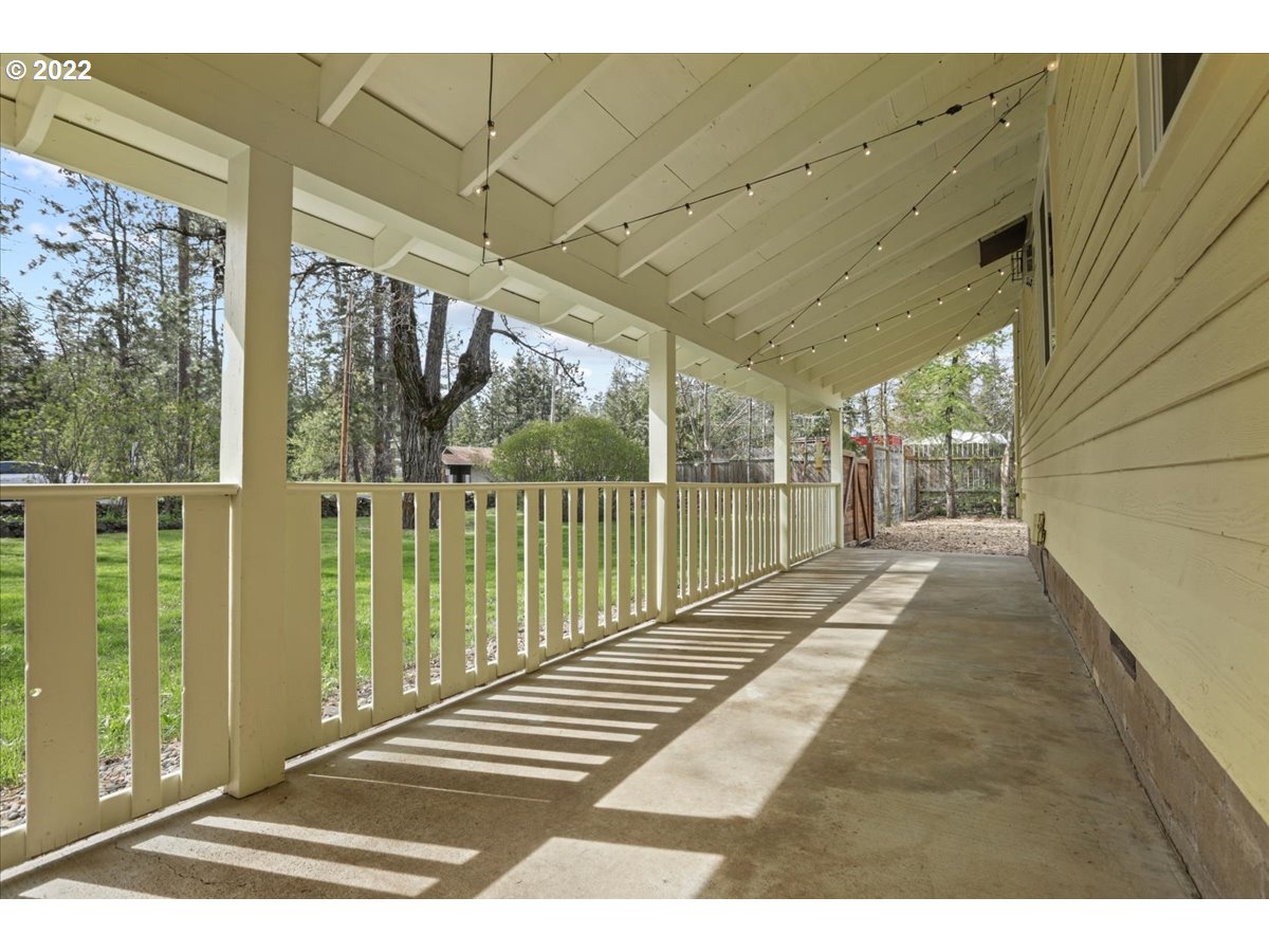 Covered concrete porch with white railings, overhead string lights, and a tree-lined yard.