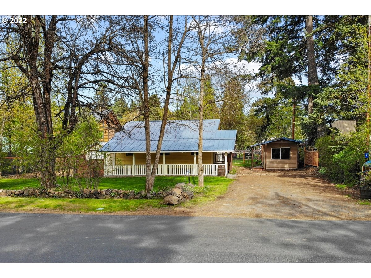 Yellow house with a white porch and metal roof, framed by trees beside a gravel driveway.