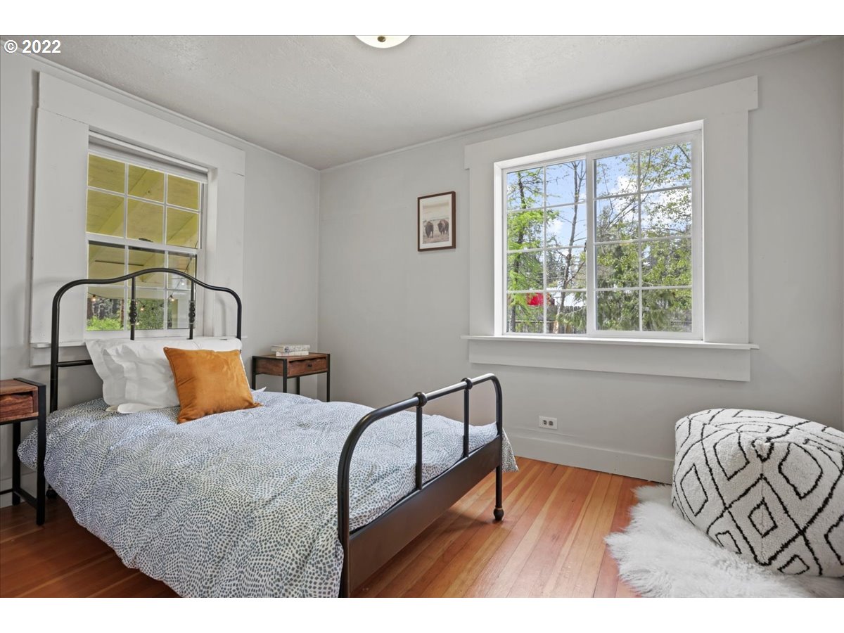 Bedroom with a black metal bed, orange accent pillow, wood floors, and large windows.