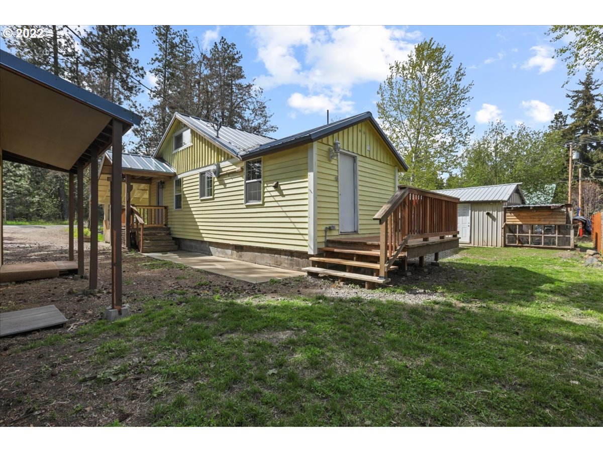 Pale yellow house with a metal roof, raised wooden porch, covered carport, and outbuildings.