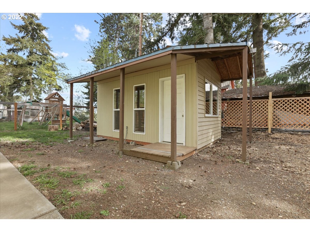 Pale-yellow outbuilding with a wide covered porch in a wooded yard.
