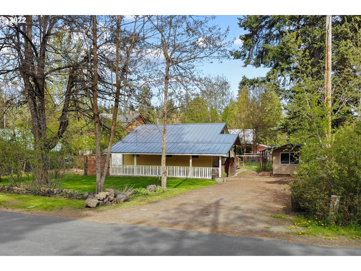 Yellow house with a metal roof and covered porch, surrounded by trees beside a gravel driveway.