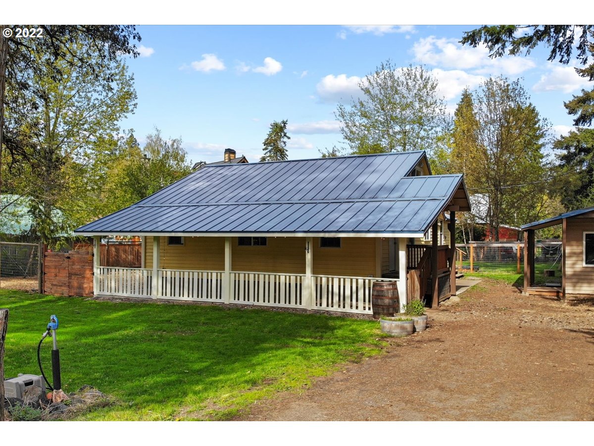 Single-story home with a broad covered porch, dark metal roof, and white railing in a wooded yard.