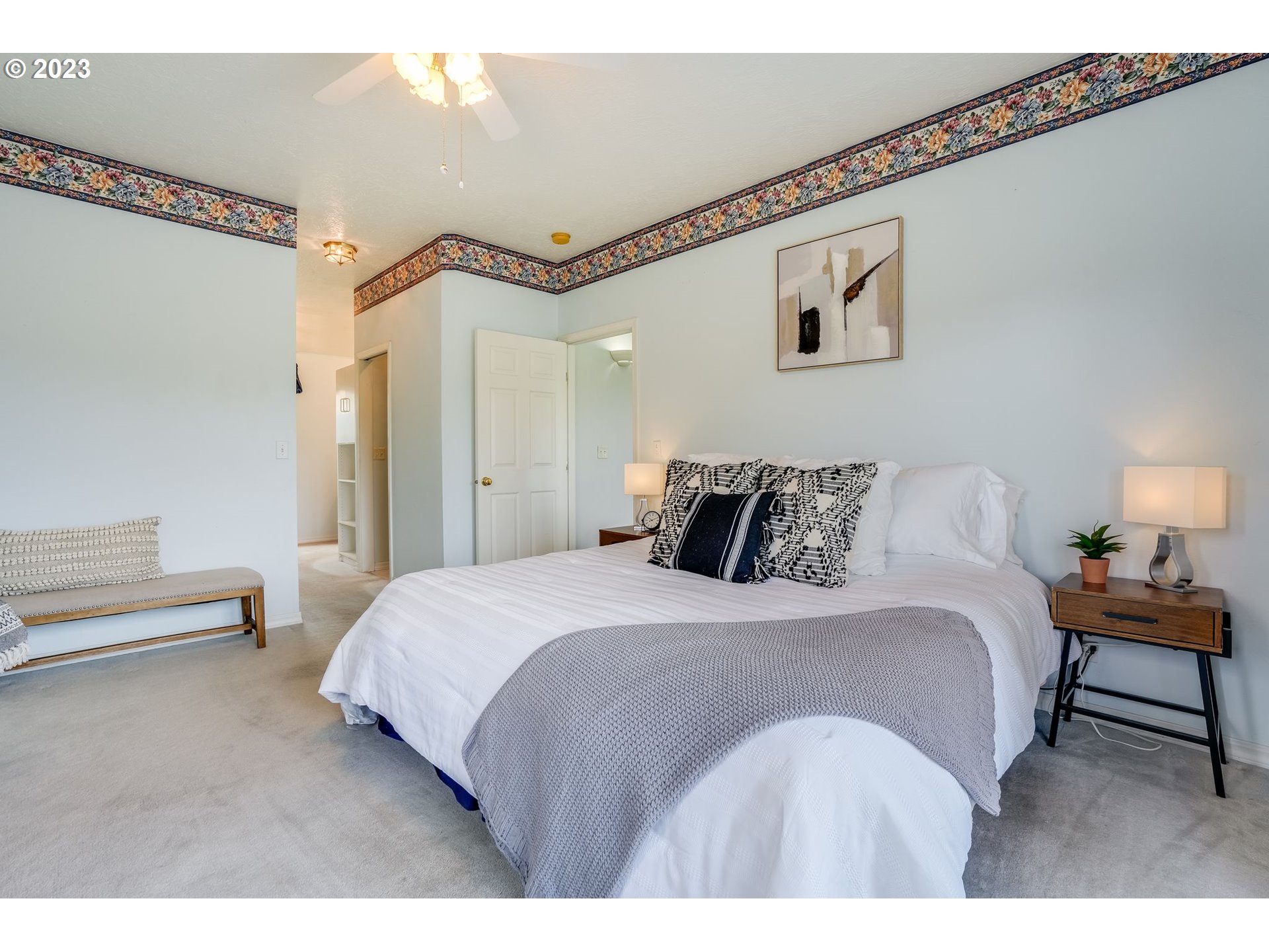 Bedroom with a white bed, gray throw, floral wallpaper border, and cushioned bench.