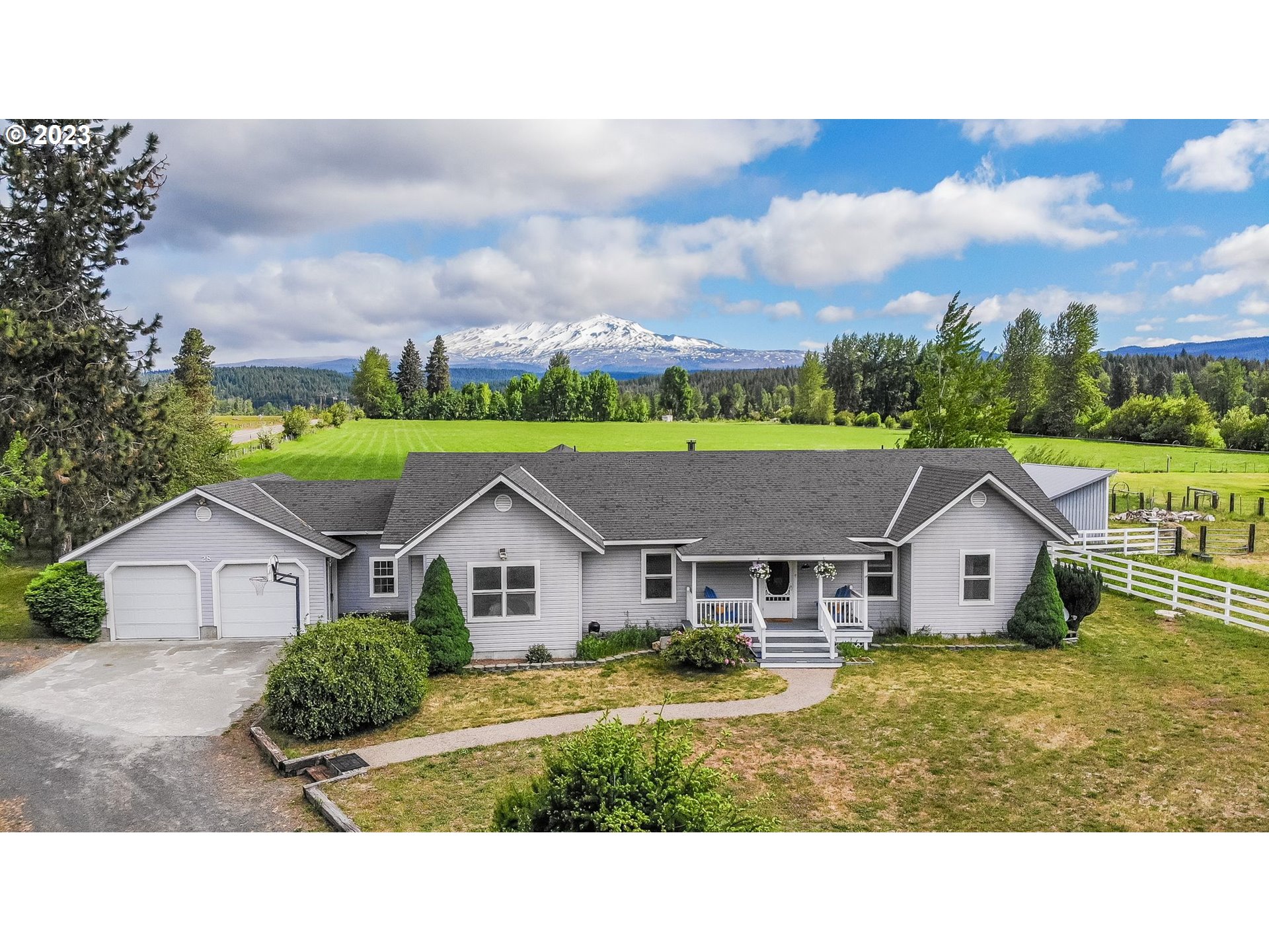 Gray single-story home with a two-car garage, backed by green fields and a snow-capped mountain.