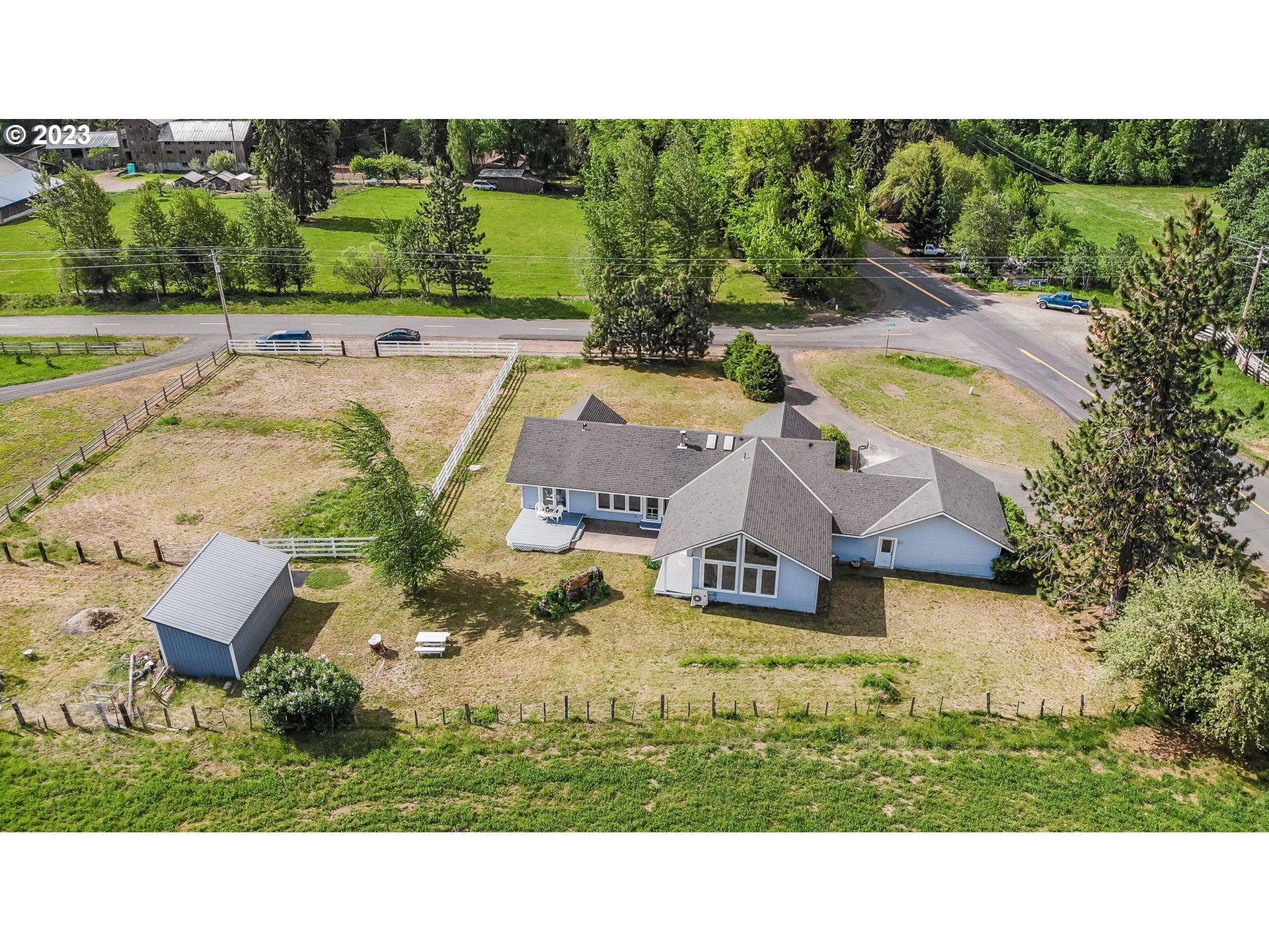 Aerial view of a gray-roofed home with a fenced yard and detached shed beside a road intersection.