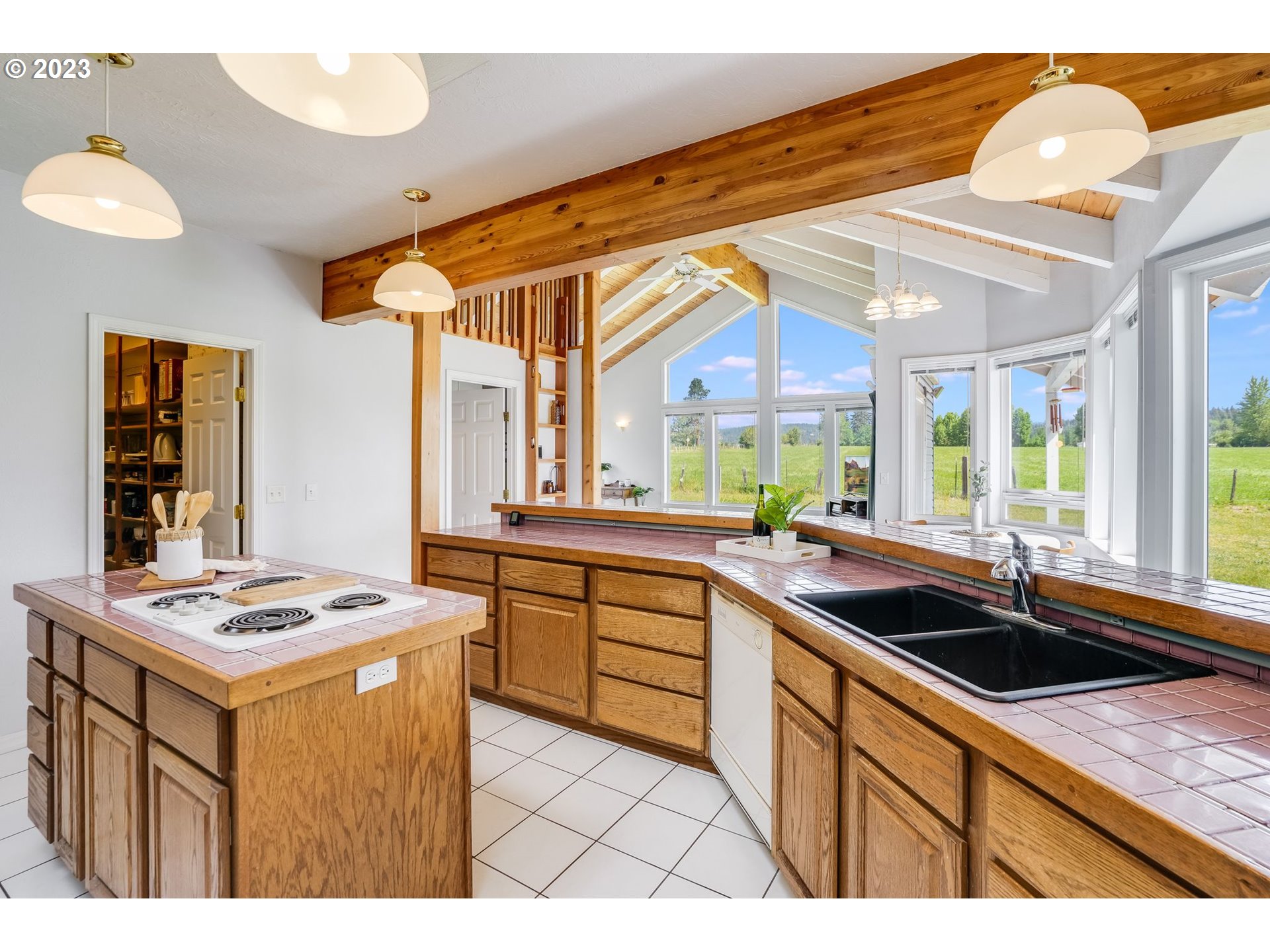 Kitchen with wood cabinetry, pink tile counters, a cooktop island, and large windows overlooking a green field.
