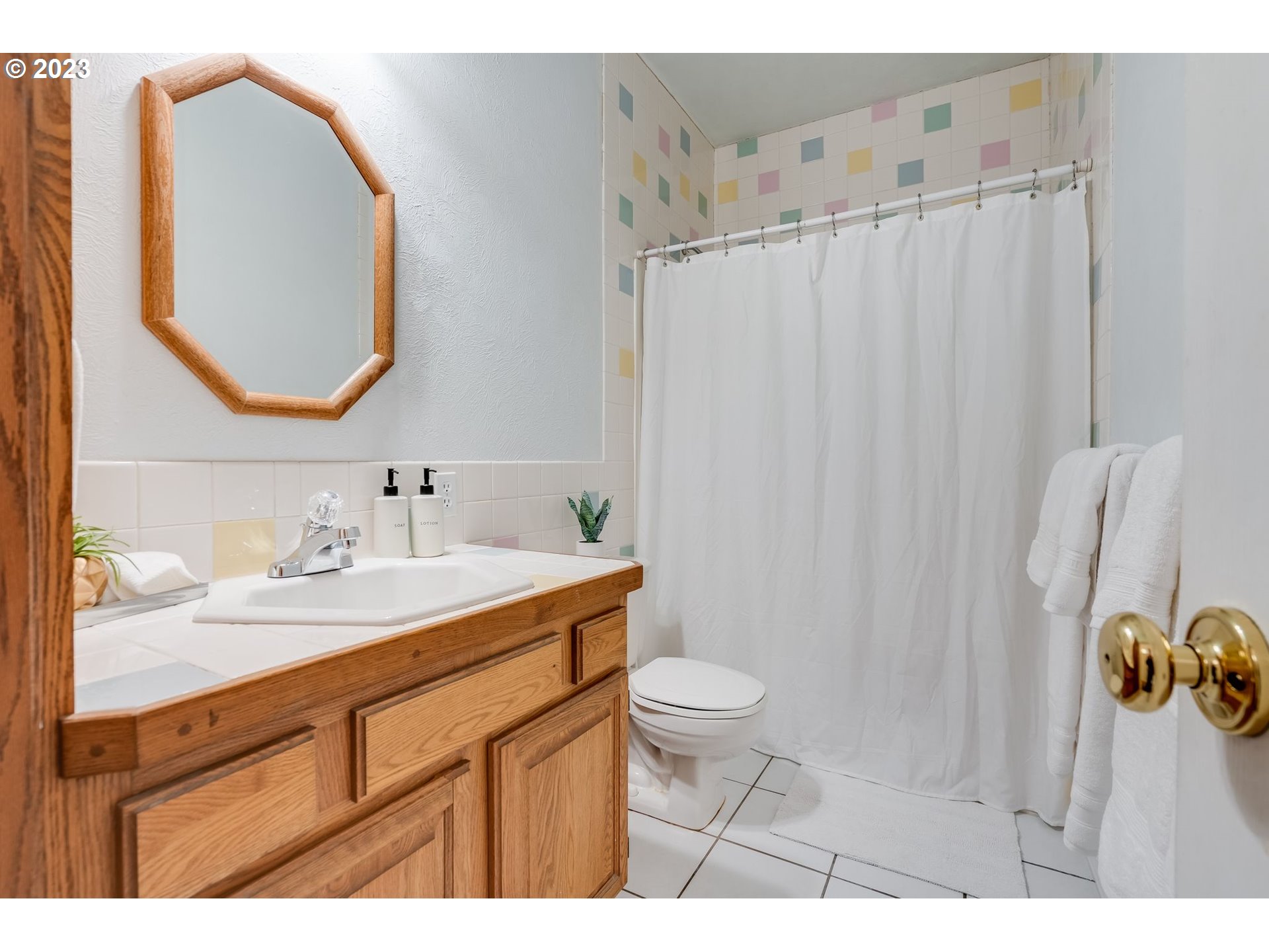 Bathroom with a wood vanity, octagonal mirror, and pastel shower tiles.