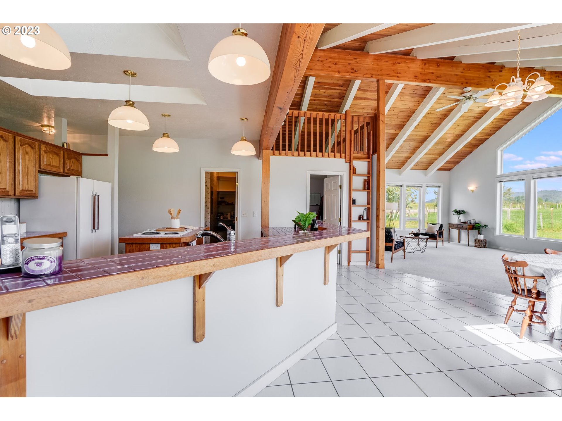 Open kitchen and living area with a tiled breakfast bar, vaulted wood ceiling, and large windows overlooking fields.