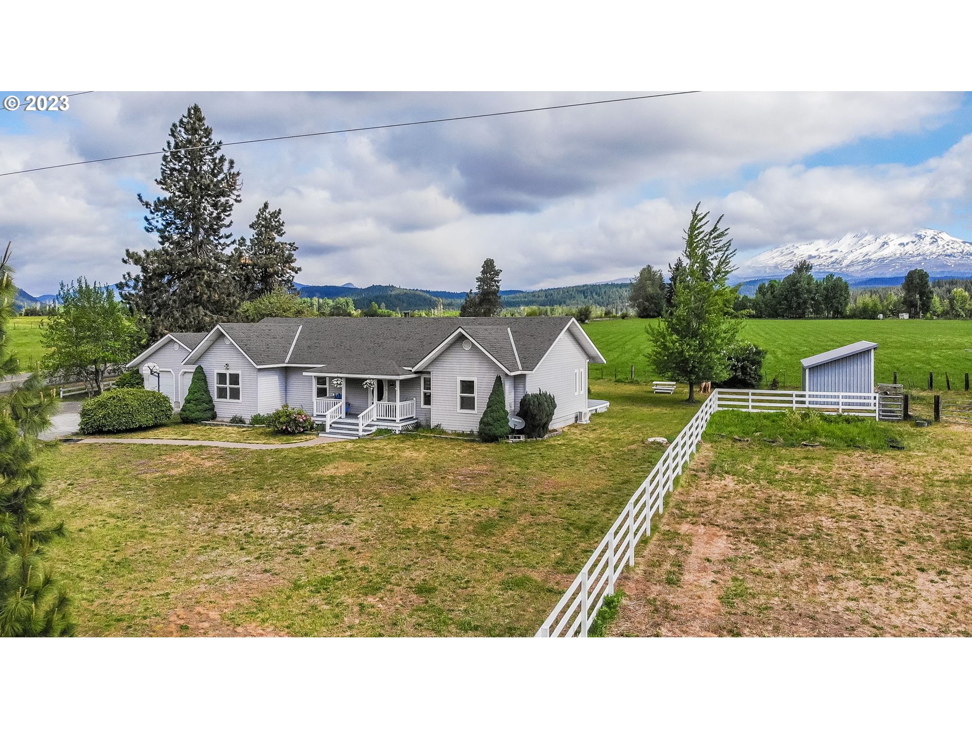 Gray ranch-style home with white fencing, pasture, outbuilding, and a snow-covered mountain beyond.