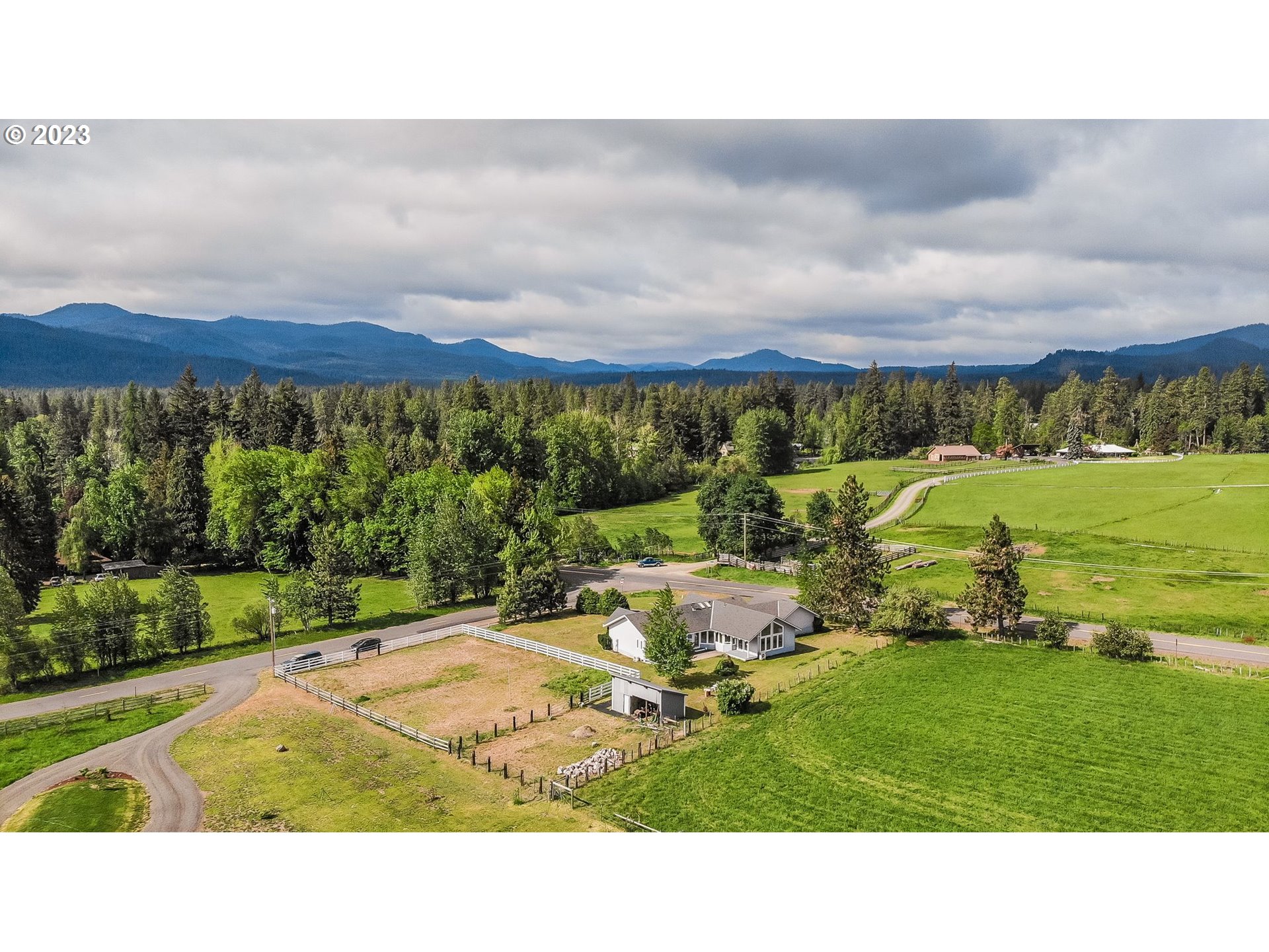Aerial view of a rural home and fenced fields, with forest and mountain ranges beyond.